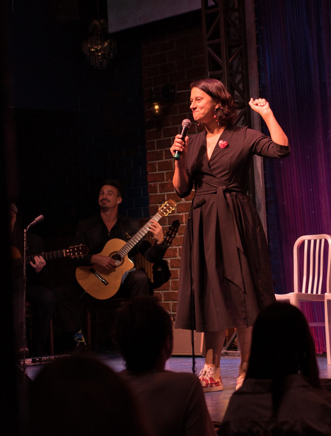 A performer in a black dress speaks into a microphone with a guitarist behind her