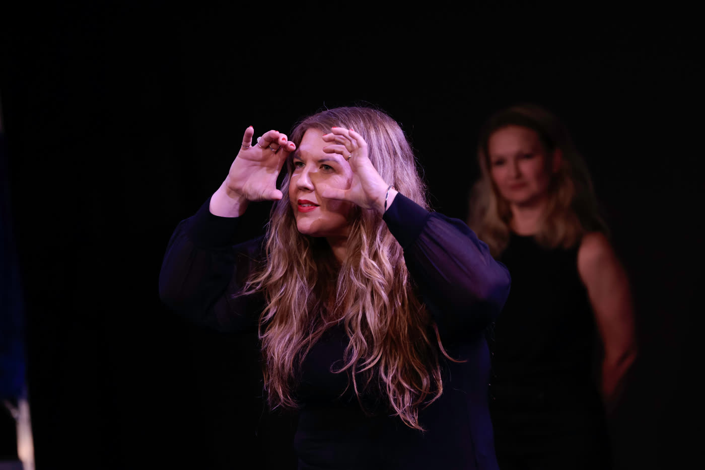 A performer framing her face with both hands under warm stage light, another performer softly out of focus behind her