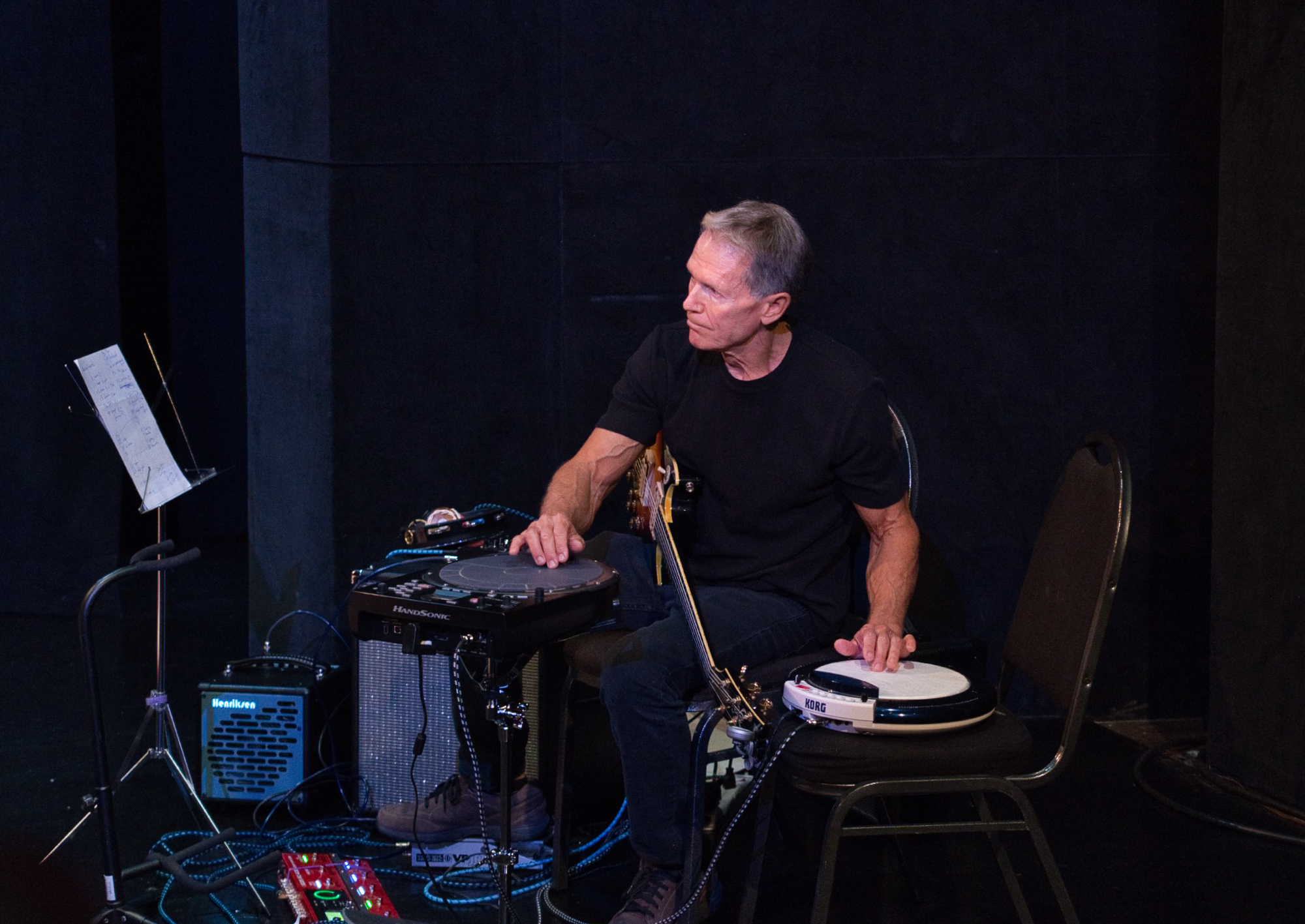 A musician playing guitar and hand percussion on a dark stage