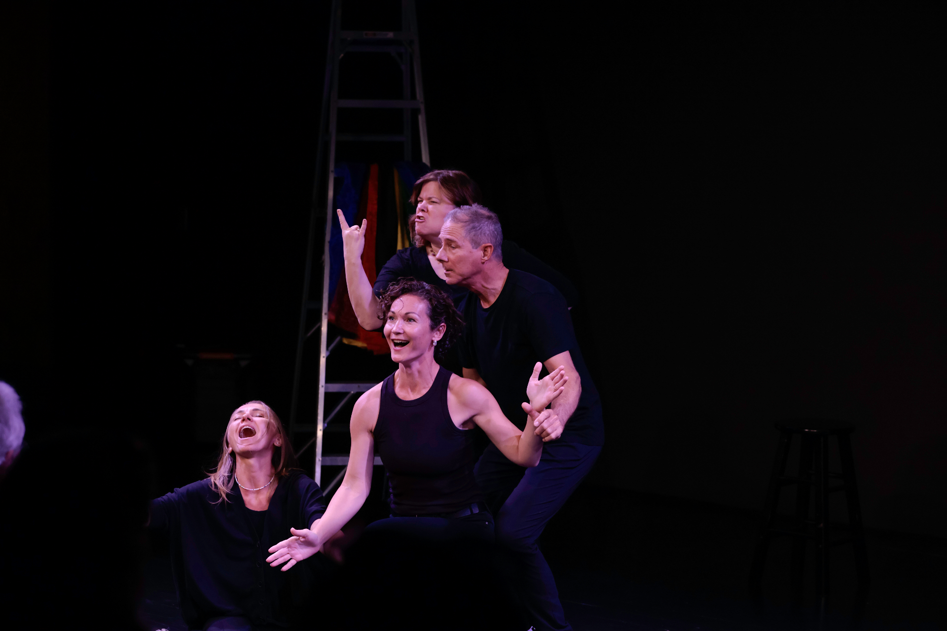 Four performers clustered by a ladder on a dark stage, one laughing, one pointing, one wide-eyed