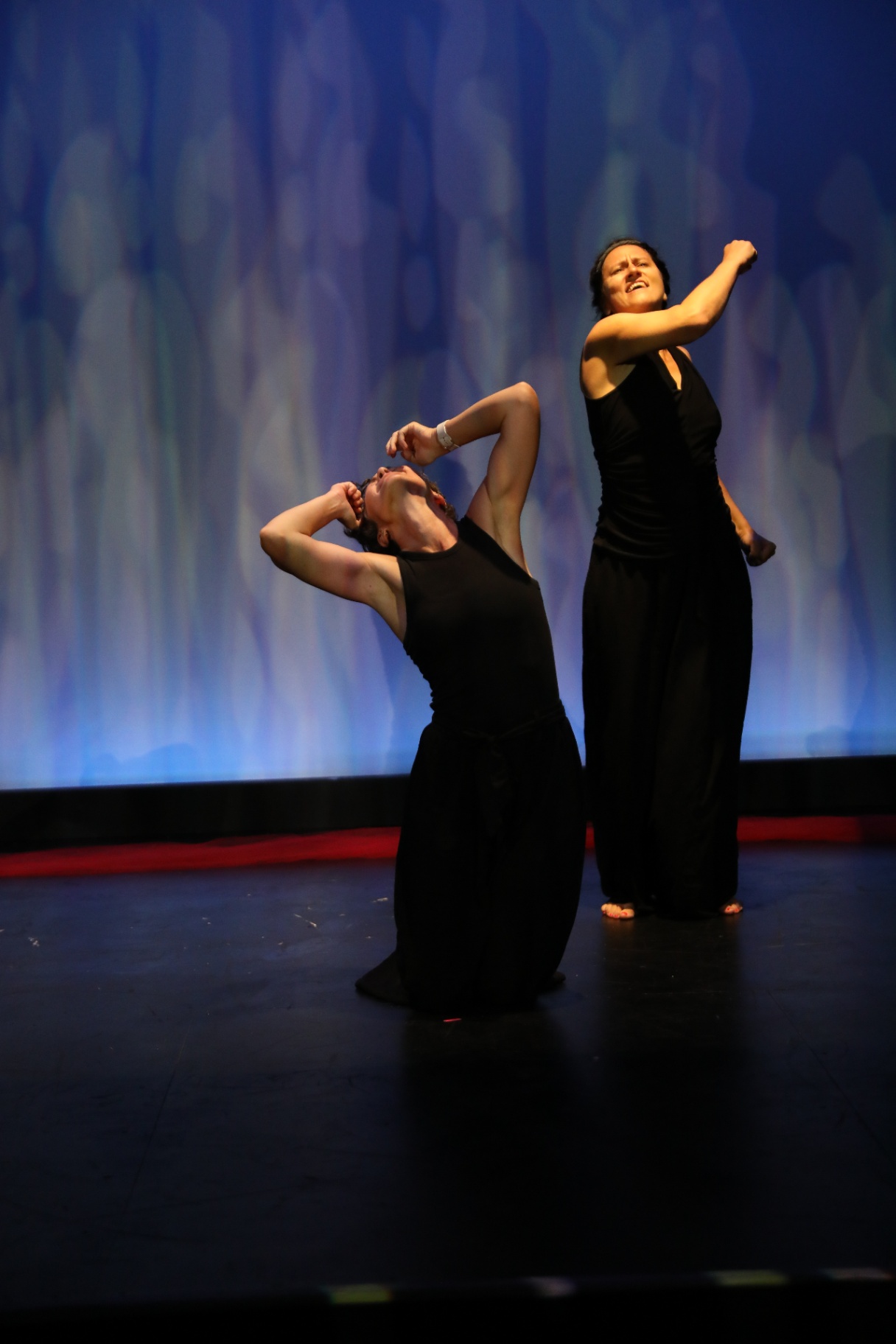 One performer kneeling in grief while another rises above her against a blue backdrop