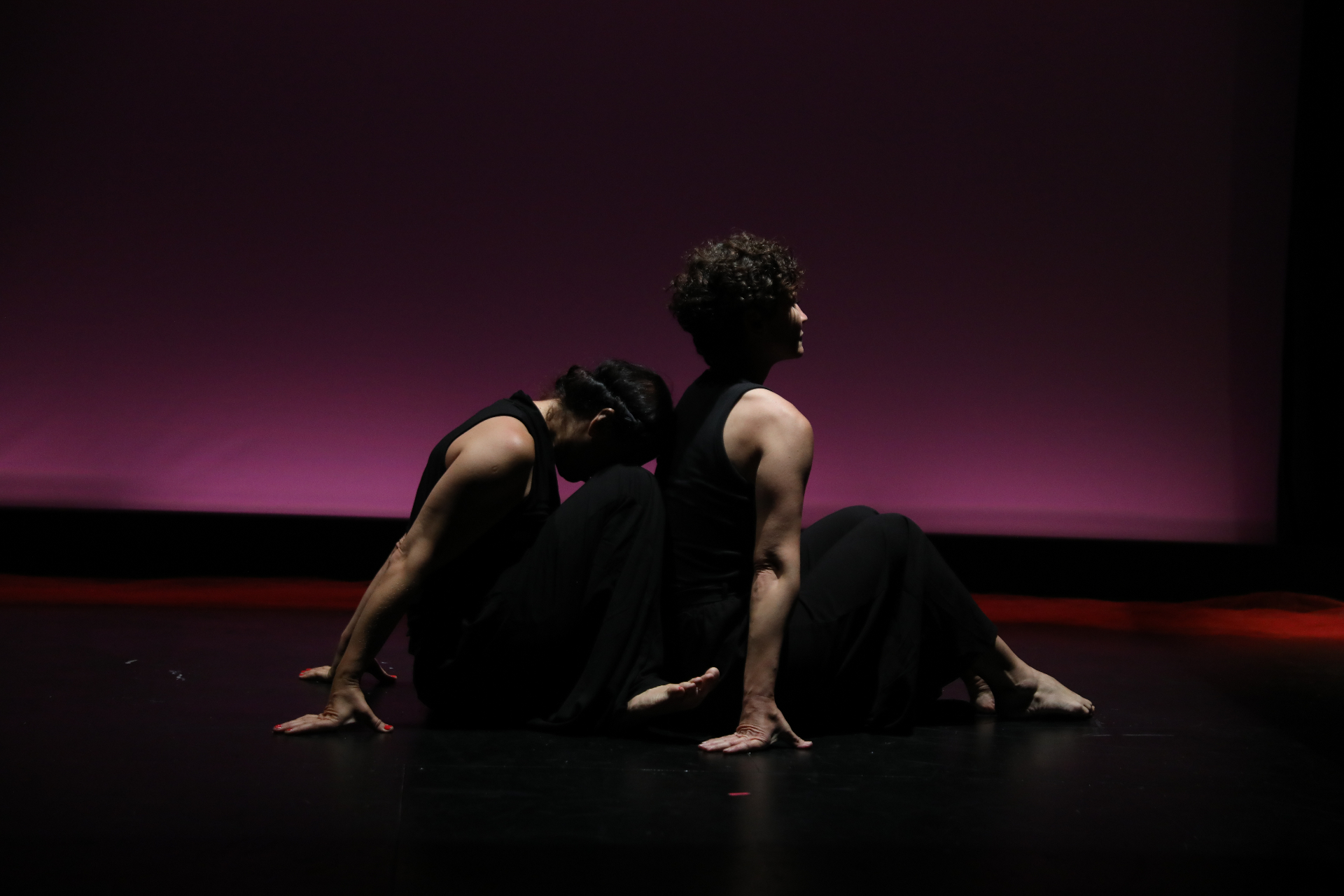Two performers seated back to back on a dark stage, quiet against a pink wash