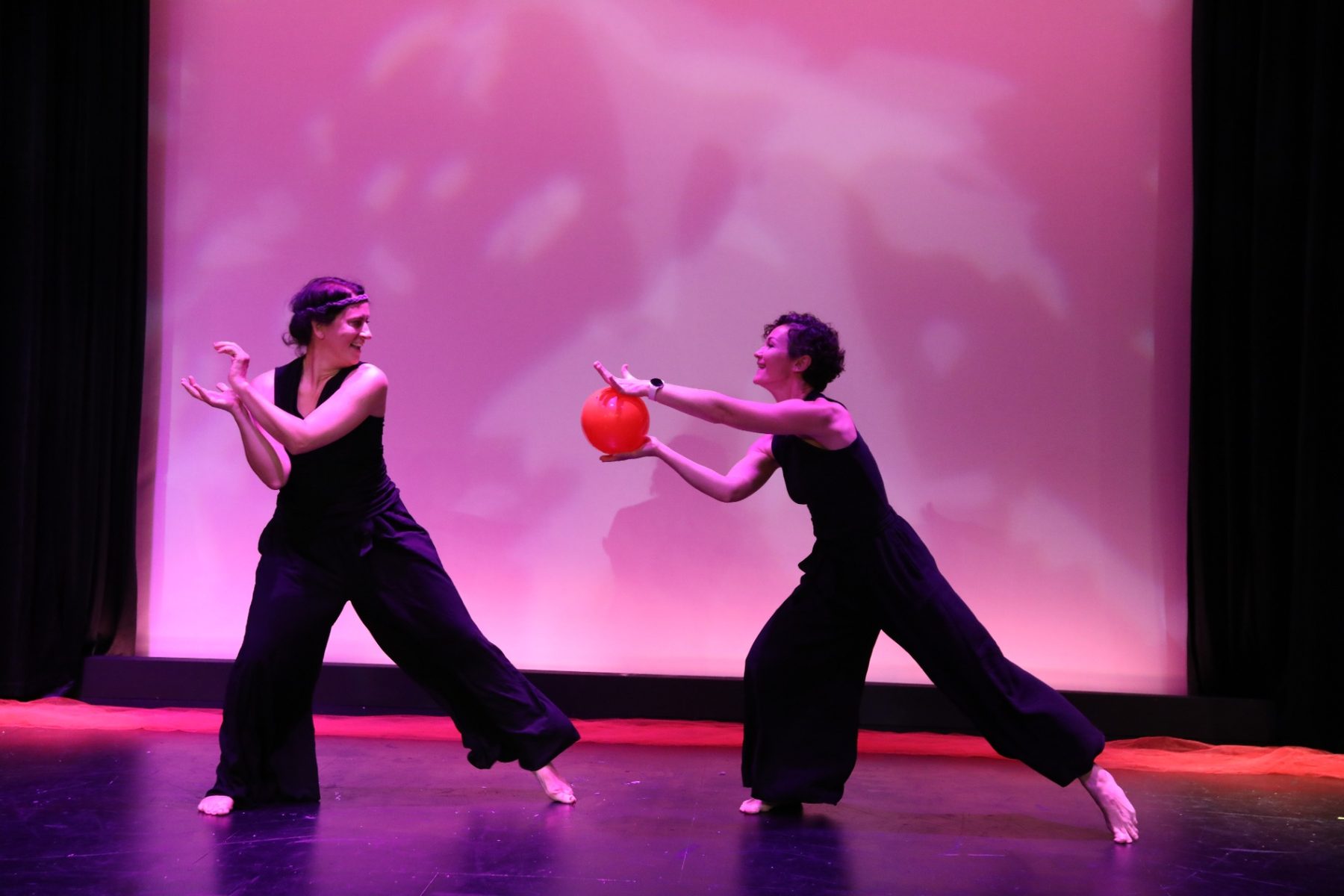 Two barefoot performers on a pink-lit stage passing a red ball